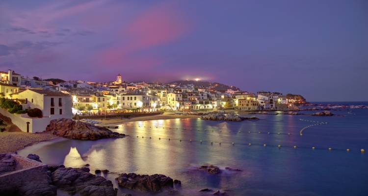 Küstenstadt in der Abenddämmerung mit beleuchteten Gebäuden und Spiegelungen auf dem Wasser.