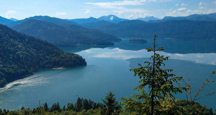 Hoher Aussichtspunkt auf einen weitläufigen blauen See, umgeben von bewaldeten Hügeln und fernen Gipfeln.