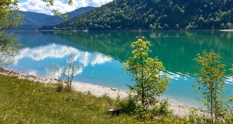 Klarer türkisfarbener See, der bewaldete Hänge unter strahlendem Sonnenschein widerspiegelt.