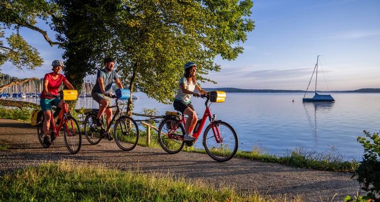 Radfahrer fahren entlang des ruhigen Seeufers zur goldenen Stunde, während Segel und Spiegelungen glitzern.