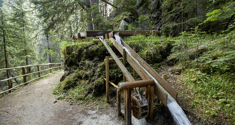 Hölzernes Wasserrad und Tröge in einer Waldumgebung.
