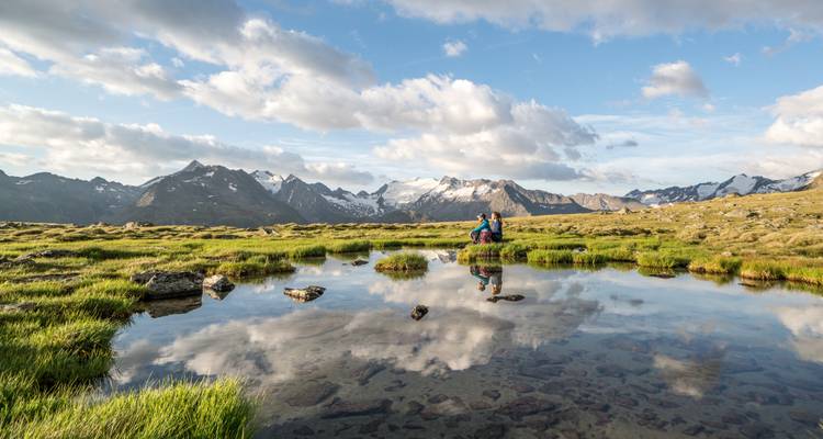 Berglandschaft mit einem spiegelnden Teich, ein Wanderer sitzt am Wasser.
