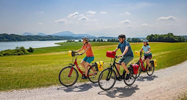 Drei Radfahrer fahren entlang eines Kieswegs am Seeufer, der von sanften grünen Feldern gesäumt ist, mit entfernten Bergen über dem Wasser.