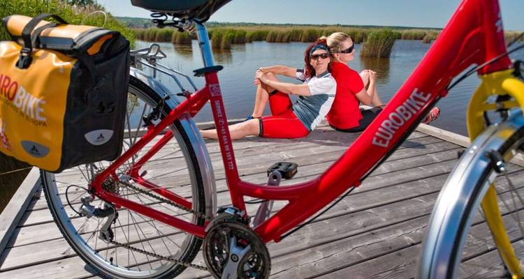 Zwei Personen sitzen neben einem geparkten Fahrrad auf einem Holzsteg in der Nähe von Wasser.