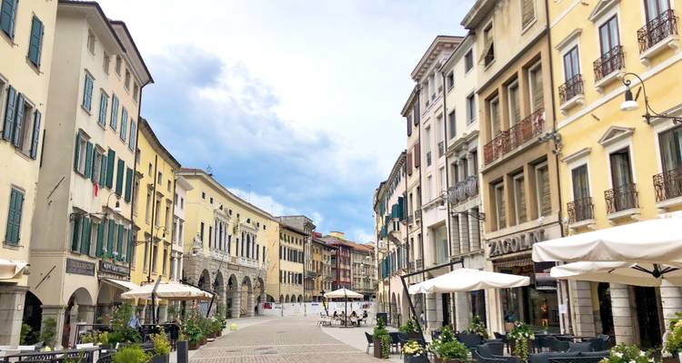 Charmante italienische Straße gesäumt von historischen pastellfarbenen Gebäuden, Arkaden und Straßencafés unter einem teilweise bewölkten Himmel.