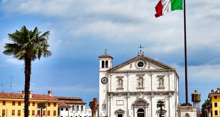 Ein italienischer Platz mit einer weißen neoklassizistischen Kirche, einem hohen Fahnenmast mit der Trikolore und einer einzelnen Palme unter dramatischen Wolken.