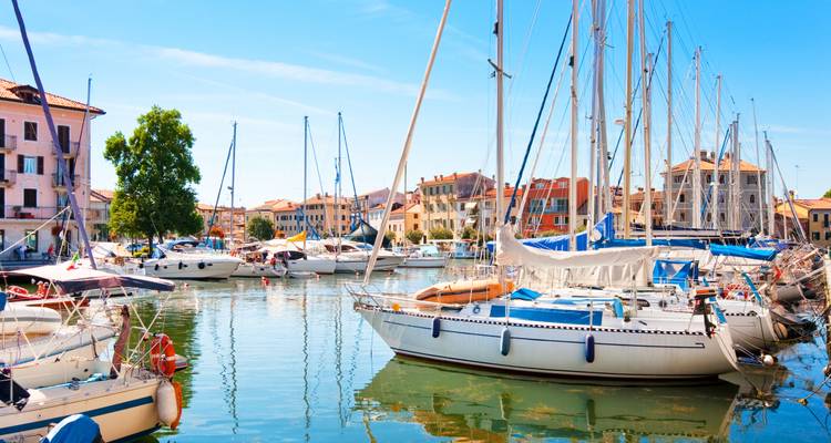Sonniger Yachthafen voller Segelboote, die sich im ruhigen Wasser einer farbenfrohen italienischen Küstenstadt spiegeln.