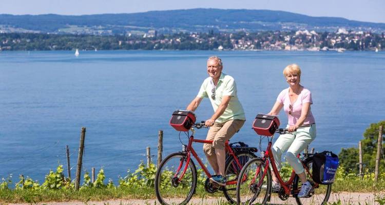 Zwei Personen fahren mit dem Fahrrad auf einem Weg entlang eines Sees mit Bergen im Hintergrund.