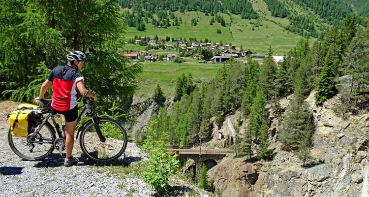 Cycliste faisant une pause sur un sentier surplombant un village avec des montagnes en arrière-plan.