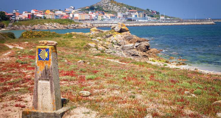 Ein Pilgermarkierungsstein mit einer Küstenstadt und dem Ozean im Hintergrund.