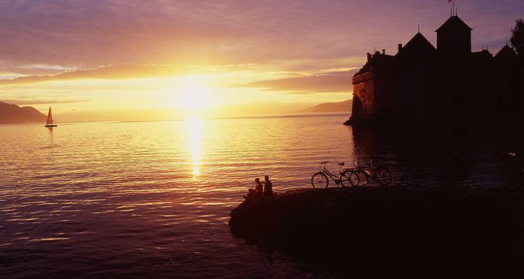 Sonnenuntergang über einem See mit silhouettiertem Schloss und Segelboot in der Ferne.