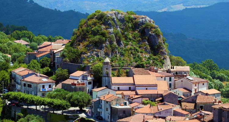 Italienisches Dorf auf einem Hügel, das sich um einen felsigen Kalksteinmonolithen gruppiert, mit Bergen im Hintergrund.
