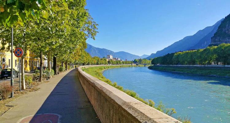 Un sendero ciclista junto al río bordeado de árboles conduce hacia un puente y edificios de colores pastel, situado entre montañas verdes bajo un cielo despejado.