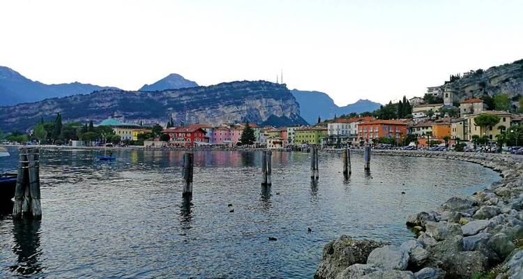 Edificios coloridos frente al agua bordean el Lago de Garda con montañas detrás y patos nadando cerca de postes de madera en el agua.