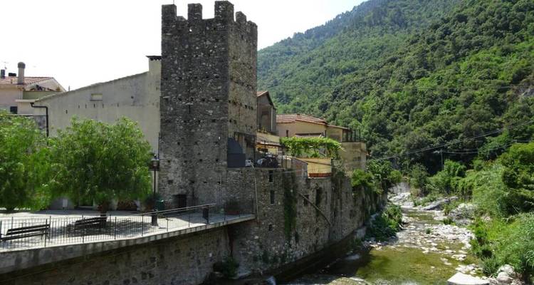 Medieval stone tower and village buildings perched beside a narrow river and forested hillside.