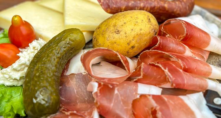 Assiette rustique avec speck, fromage, pomme de terre, cornichon et légumes frais sur une planche en bois.