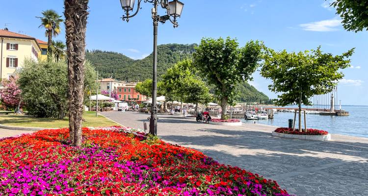 Des parterres de fleurs colorés bordant une promenade au bord du lac avec des cafés, des bateaux et des collines lointaines sur le lac de Garde.