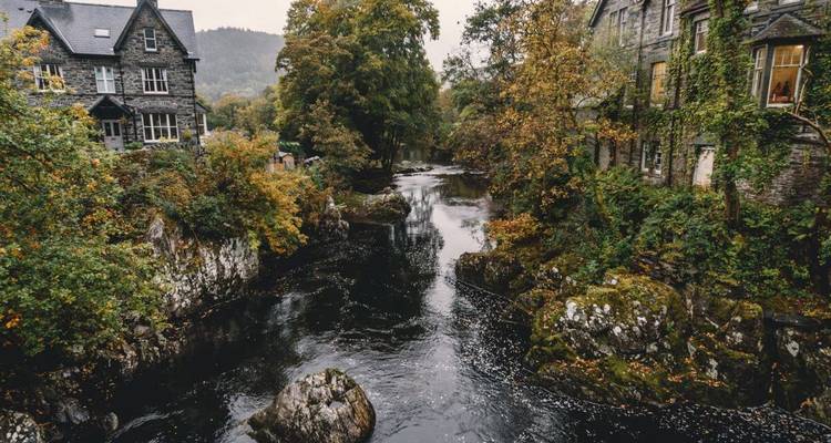 Malerischer Fluss, der zwischen Steingebäuden und üppigen Herbstbäumen in einer walisischen Dorflandschaft fließt