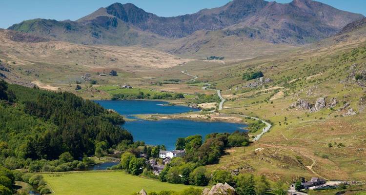 Breites Tal mit gewundener Straße, Bäumen und zwei Seen unter rauen Gipfeln in Snowdonia.