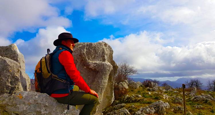 Wanderer mit Rucksack und Hut, der auf einem Kalksteinfelsen sitzt und die weitläufigen Bergpanoramen unter blauem Himmel genießt.