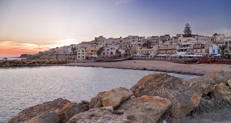 Friedlicher Strand mit felsiger Küste und Gebäuden bei Sonnenuntergang.