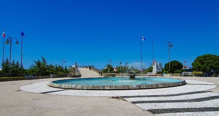 Offener Platz mit einem modernen kreisförmigen Brunnen, Flaggen und einer Rampe, die zu einer Küstenpromenade unter einem strahlend blauen Himmel führt.