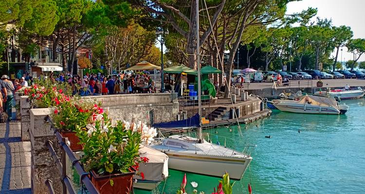 Paseo bullicioso junto al lago bordeado de puestos de mercado, flores y embarcaciones de recreo en aguas turquesas.