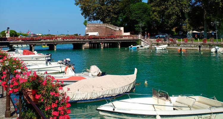 Vista enmarcada por flores de pequeñas embarcaciones de recreo amarradas a lo largo de un canal turquesa bajo un puente de piedra.