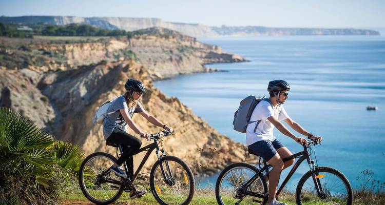 Deux personnes faisant du vélo sur un sentier côtier avec des falaises et la mer en arrière-plan.