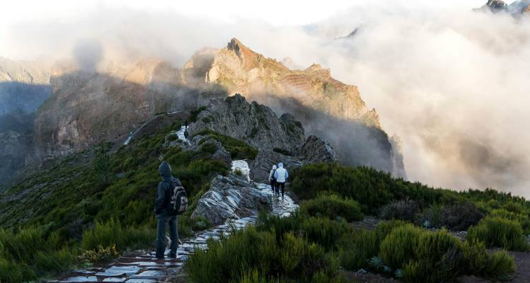 Wandelaars op een rotsachtig bergpad met wolken en bergtoppen.