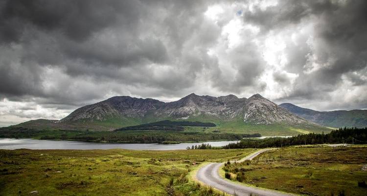 Des nuages orageux planent au-dessus de montagnes escarpées et d'une route rurale sinueuse bordant un lac.