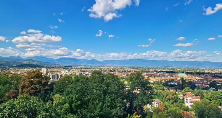 Amplia vista panorámica sobre una ciudad italiana con techos de terracota enmarcada por colinas verdes y montañas distantes bajo nubes dispersas.