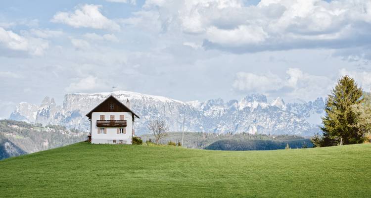 Isolated white alpine house on a vivid green meadow with dramatic snow-clad Dolomite peaks in the background and a partly cloudy sky.