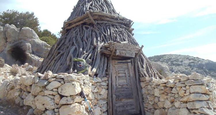 Traditionelle, kegelförmige Schäferhütte aus Treibholzstämmen, umgeben von einer niedrigen Steinmauer in unwegsamem Gelände.
