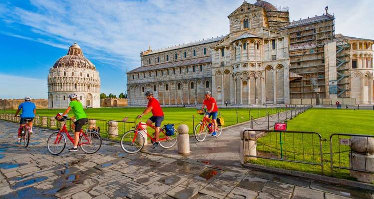 Cyclists ride past Pisa's Cathedral, Baptistery and Leaning Tower on a bright day.