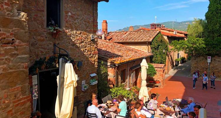 Outdoor terrace of a stone village restaurant in Tuscany where travellers enjoy food under umbrellas.