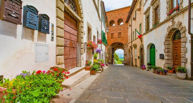 Charming cobblestoned lane in an Italian hill town framed by stone houses, colorful flowers, and an arched brick gate at the end.