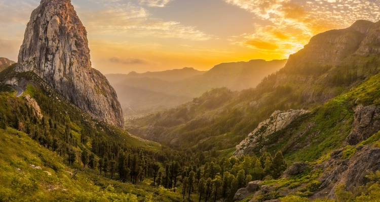 Dramatic sunset over lush volcanic valley with towering rock monolith and winding road.