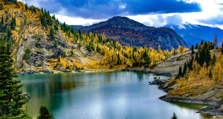 Ein ruhiger See, umgeben von bunten Herbstbäumen und Bergen in der Ferne.