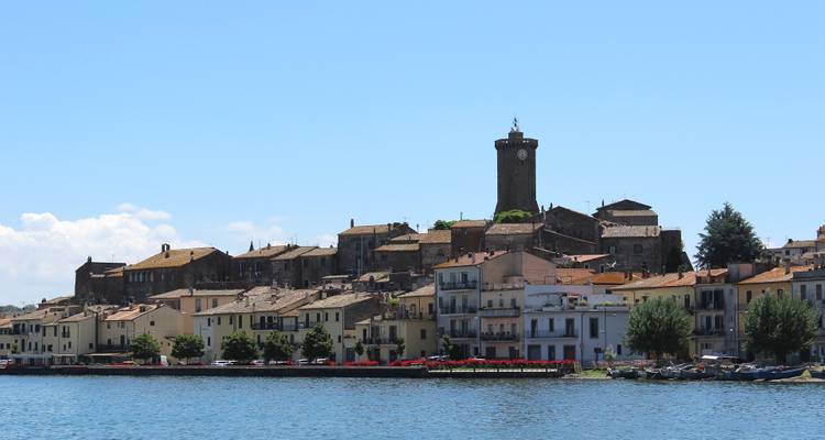 Vista a orillas del agua de la ciudad de Bolsena con edificios en tonos pastel y una torre de reloj de piedra que se eleva detrás de la costa del lago.