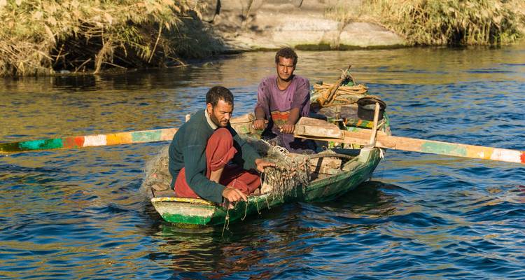 Two fishermen on a small boat in a river with green banks.