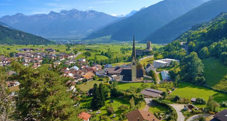 Malerisches Alpendorf mit hohem Kirchturm in grünem Tal unterhalb schneebedeckter Gipfel.