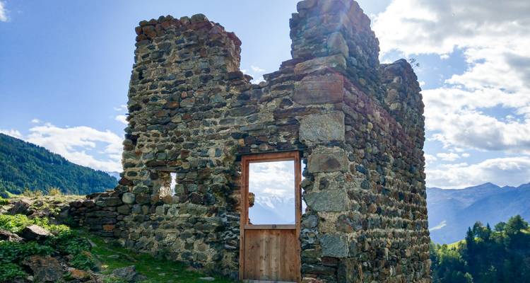 Eine verwitterte Steinruine steht auf einem grasbewachsenen Bergrücken mit fernen Alpengipfeln unter einem strahlend blauen Himmel.