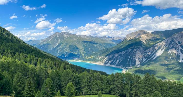 Ein leuchtend türkisfarbener See schmiegt sich zwischen dichte Wälder und aufragende Alpengipfel unter einem hellen Himmel voller flauschiger Wolken.