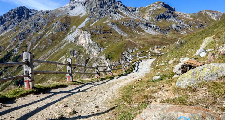 Ein gewundener Bergpfad, der von einem Holzzaun gesäumt wird, führt einen zerklüfteten Hang hinauf unter klarem blauem Himmel.