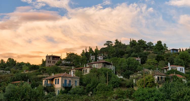 Traditional stone houses on a wooded hillside at sunset with pastel clouds overhead.