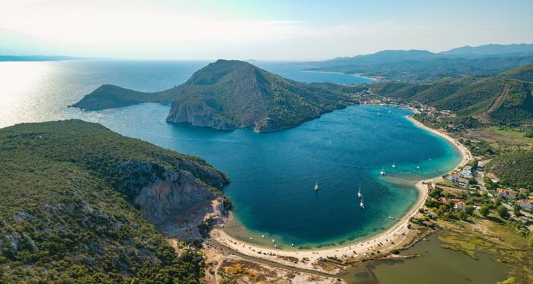 Aerial view of a turquoise bay surrounded by green headlands and sandy beaches dotted with sailboats.
