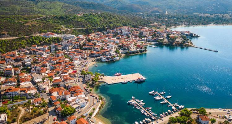 Drone shot of Neos Marmaras town with red-tiled roofs, marina, and forested hills surrounding a clear blue bay.