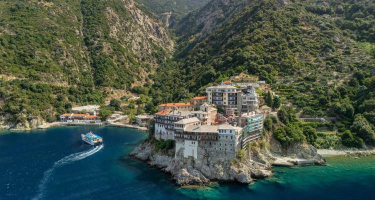 Clifftop monastery complex on Mount Athos facing deep blue waters with a ferry passing below.