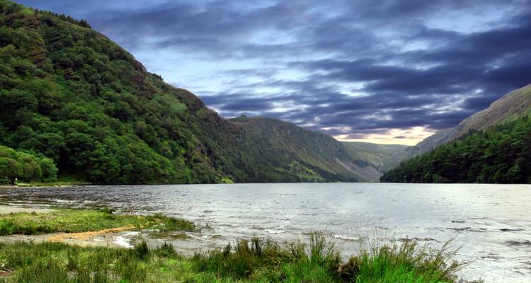 Lac entouré de montagnes verdoyantes sous un ciel dramatique.